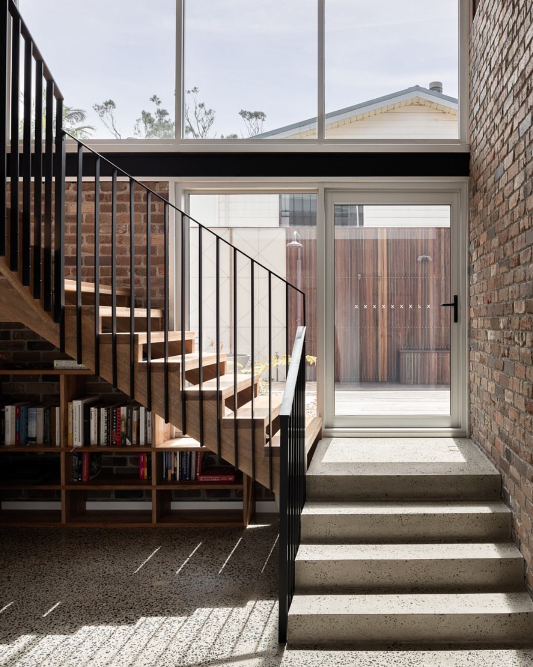 Interior of a custom residential build showing a timber and steel staircase with integrated bookshelf storage beneath, terrazzo flooring, exposed brick wall, and a glass door opening to a private courtyard with timber screening, demonstrating the craftsmanship and space-efficiency achievable in a custom studio home