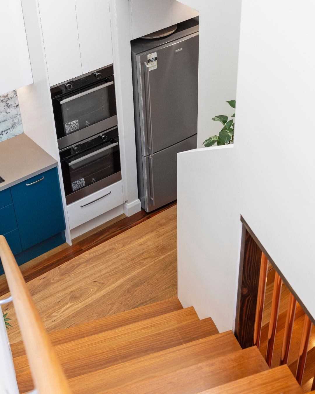 Interior of a completed custom townhouse showing a timber staircase descending past a navy blue kitchen with stainless steel appliances and warm timber flooring, demonstrating the multi-level layout and high-specification finish of a CKS Projects Illawarra townhouse build